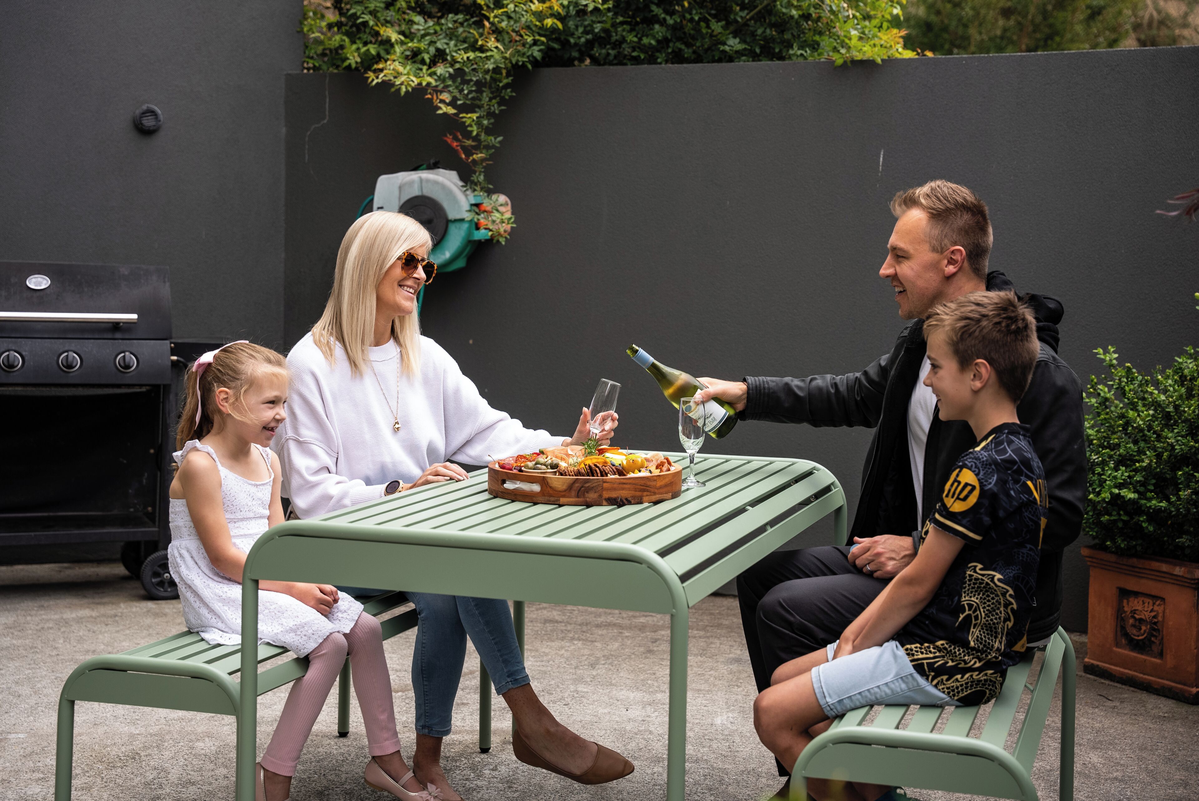 A family of four seated at an outdoor table with a grill and a potted plant in the background.