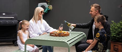 A family of four seated at an outdoor table with a grill and a potted plant in the background.
