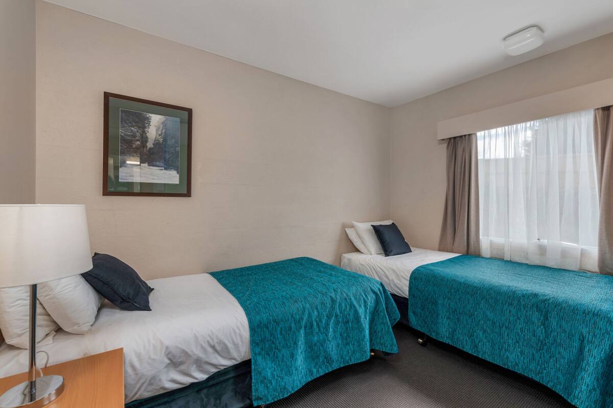 A hotel room with two single beds, a nightstand with a lamp, a framed picture on the wall, and a window with curtains.