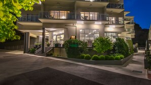A multi-story hotel with a modern design, featuring balconies on each floor, a well-lit entrance, and a landscaped garden area.