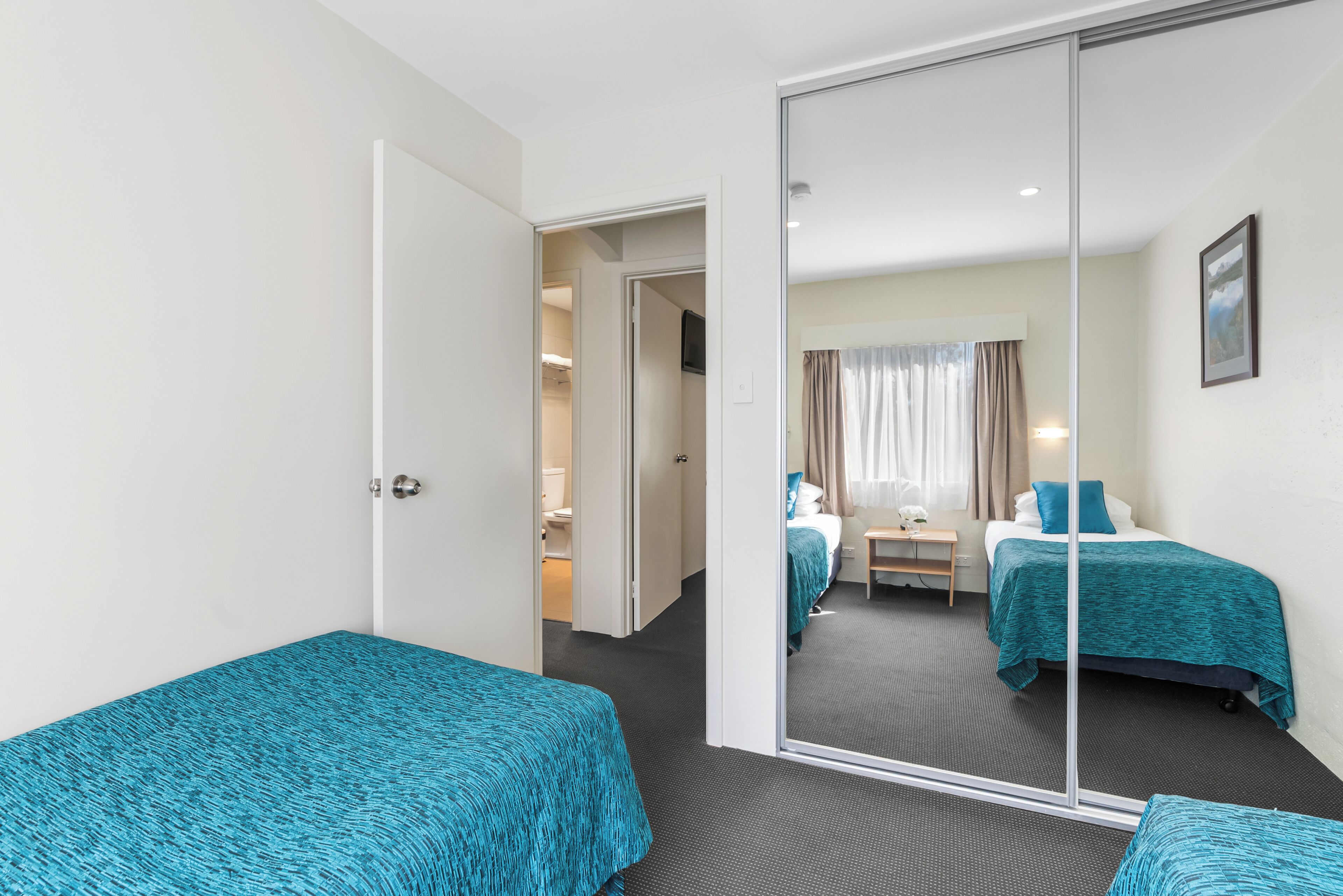 Habitación de hotel con armario con espejo, dos camas con ropa de cama color turquesa, un escritorio con silla y una ventana con cortinas.