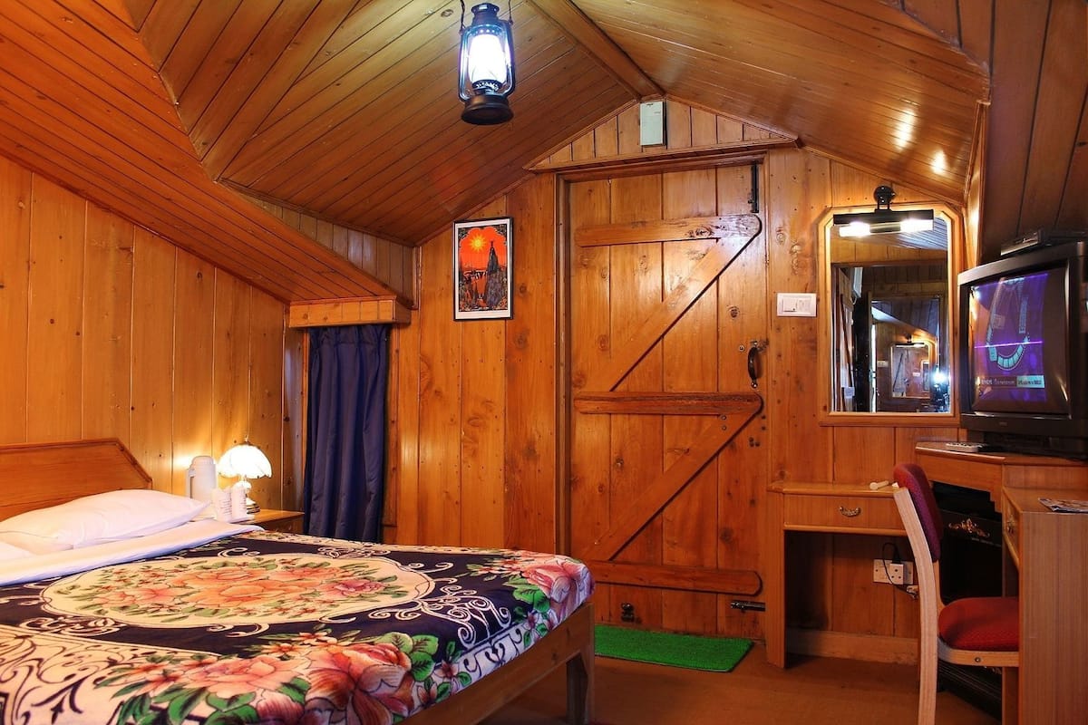 A wooden cabin room with a bed, a desk, a chair, a television, and a door.