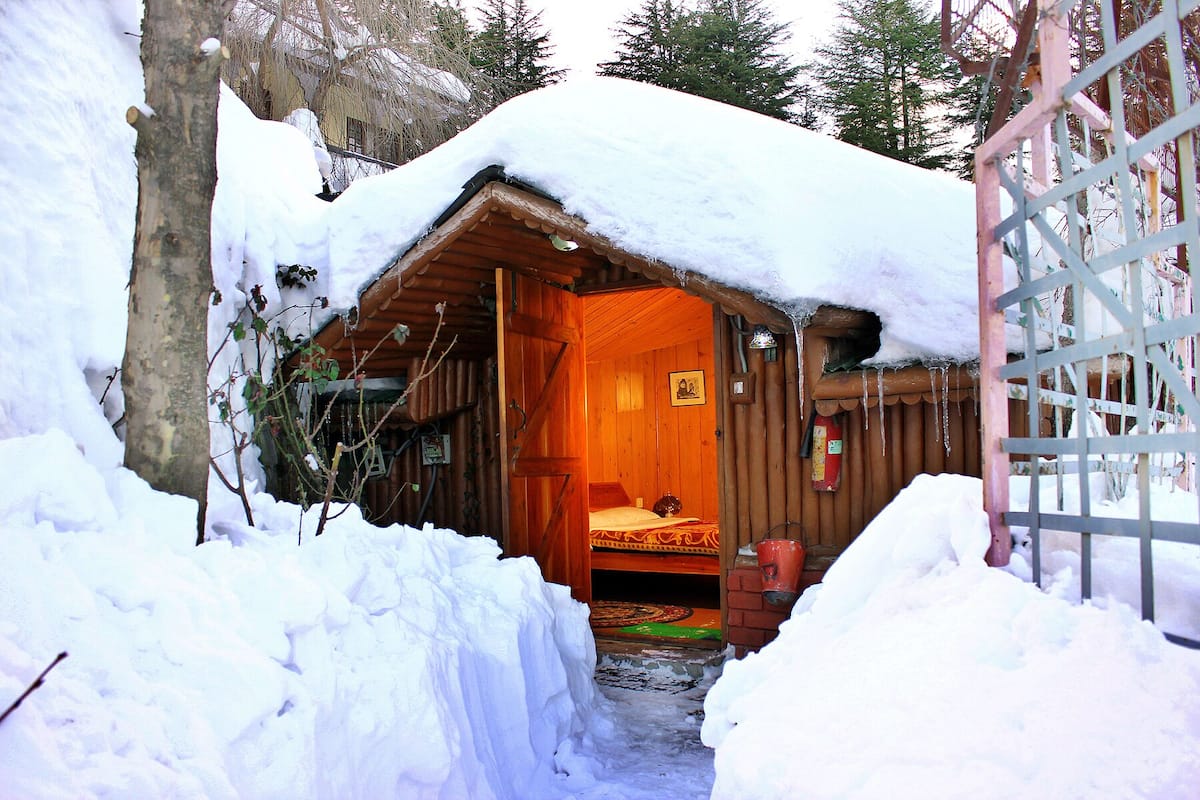 A wooden cabin with a snow-covered roof, a visible bed inside, and a small window with a picture.