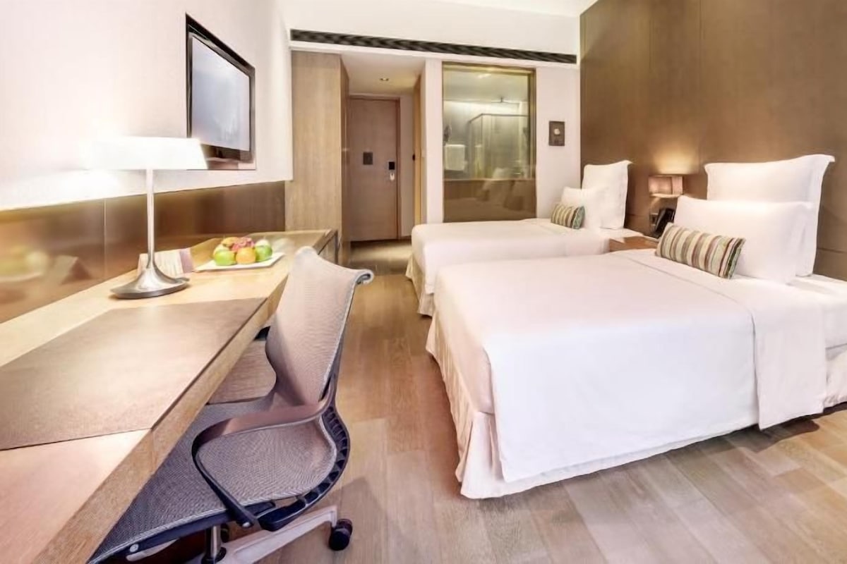 A hotel room with two beds, a desk with a chair, a TV, and a bathroom visible through an open door.