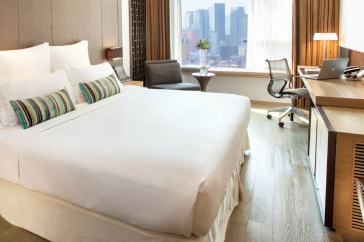 A hotel room with a large bed, a desk with a laptop, a chair, and a view of the cityscape through the window.