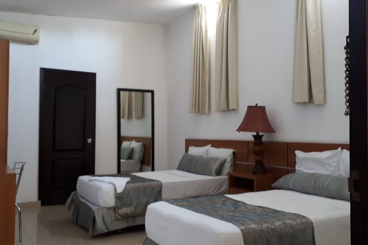 Une chambre d’hôtel avec deux lits simples, une tête de lit en bois, une table de chevet avec une lampe, un miroir et une porte.