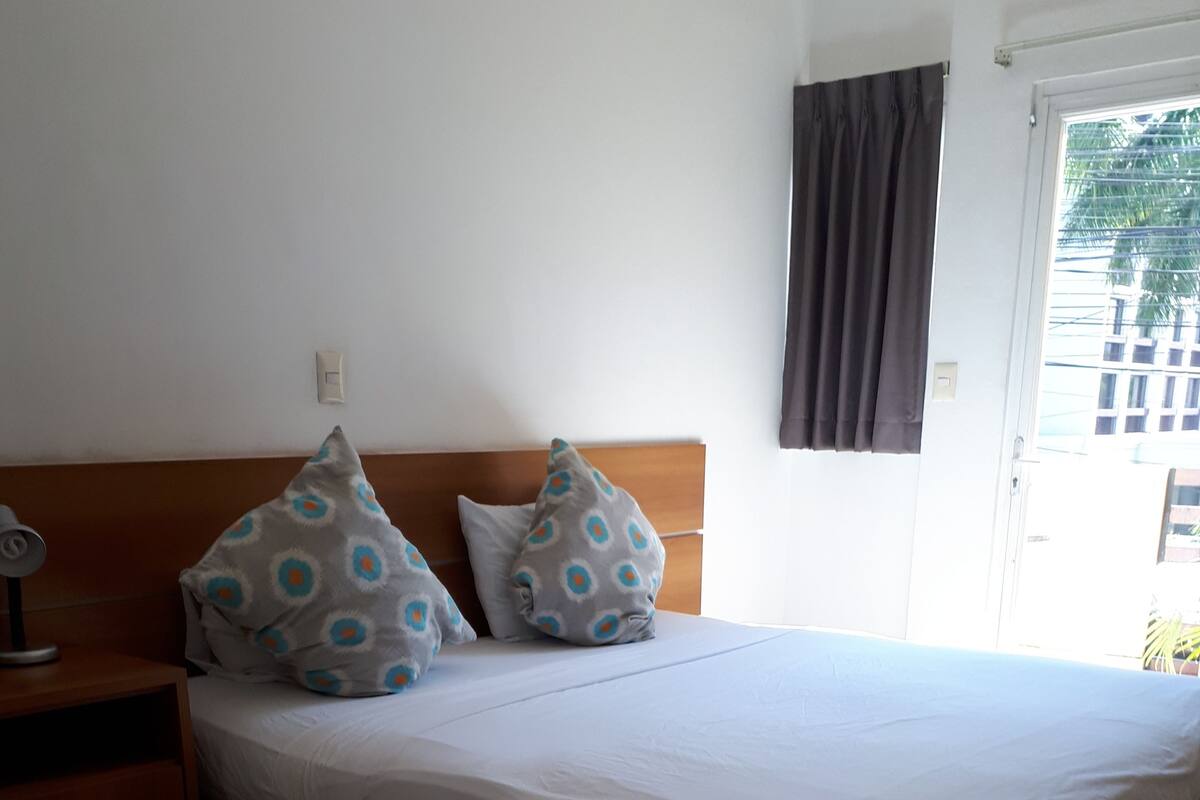 Une chambre à coucher avec un lit, deux oreillers, une table de chevet, une lampe et une fenêtre avec des rideaux.