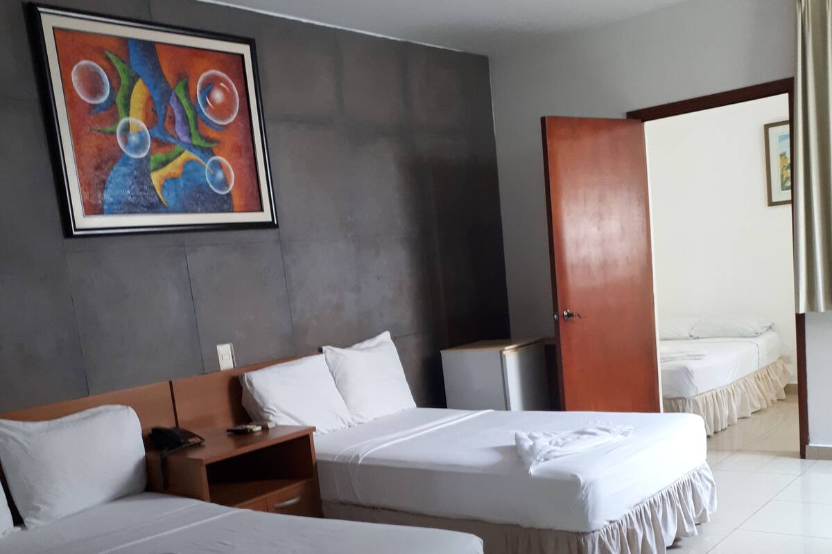 Une chambre d’hôtel avec deux lits, un bureau, une chaise, une table de chevet, un tableau au mur et une porte donnant sur une autre pièce.
