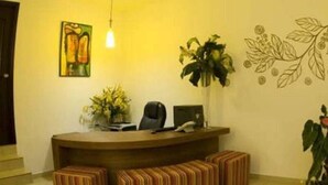 Mostrador de recepción con computadora, una silla y una planta. Hay bancos a rayas frente al mostrador.
