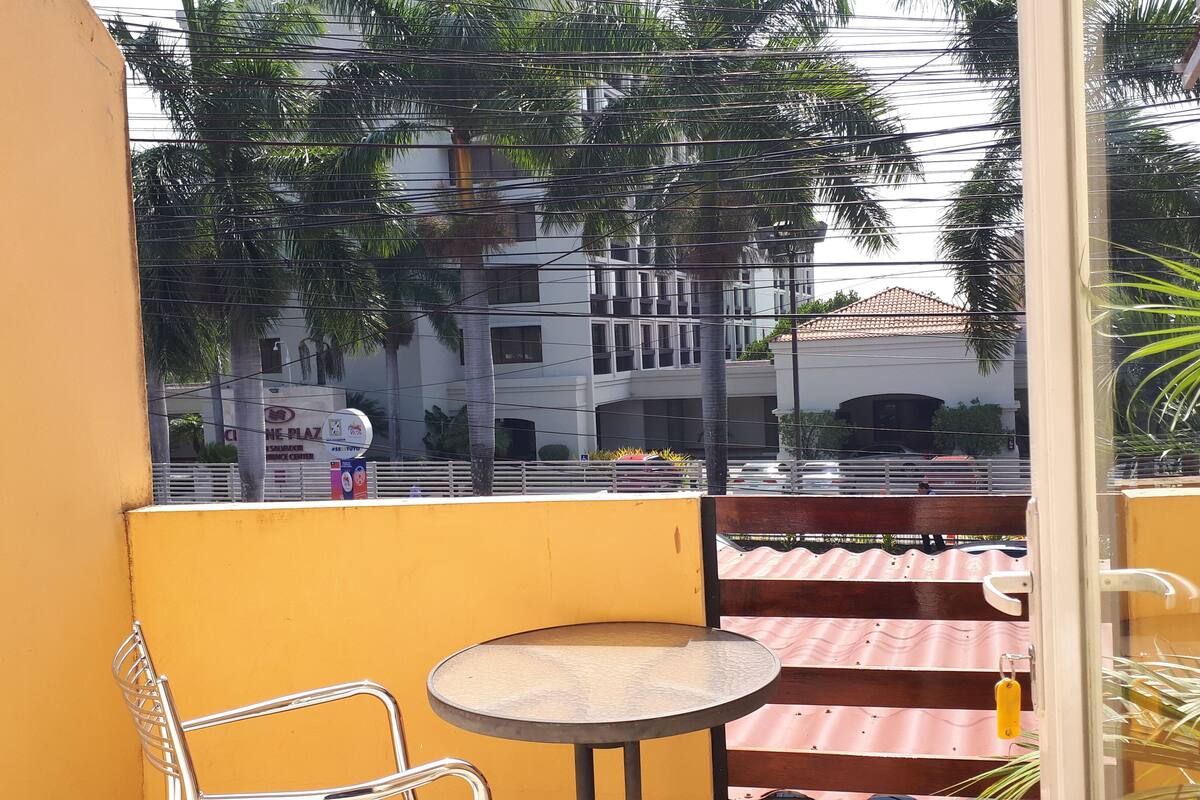 Un balcon avec un mur jaune, une table ronde et une chaise en métal. Au fond, on aperçoit un immeuble avec une enseigne indiquant « CLINIC PLAZA ».