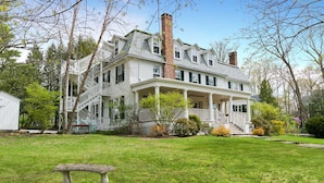 A large, two-story house with a wrap-around porch, a brick chimney, and a small garden area with a bench. - The Dewey (Lenox)