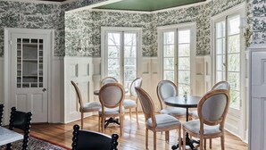 A dining room with a wooden table and chairs, large windows with white frames, and a patterned wallpaper. - The Dewey (Lenox)