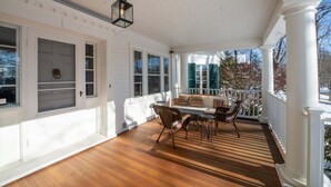 A covered porch with a wooden floor, white columns, and a seating area with chairs and a table. - The Dewey (Lenox)