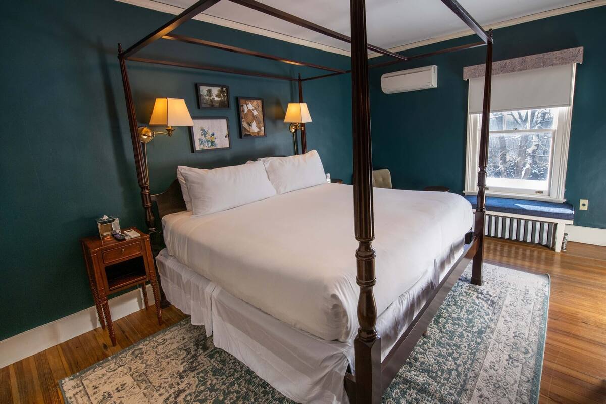 Ein Himmelbett mit weißen Leinen, ein Nachttisch mit Lampe und ein Fenster mit Blick auf Bäume.