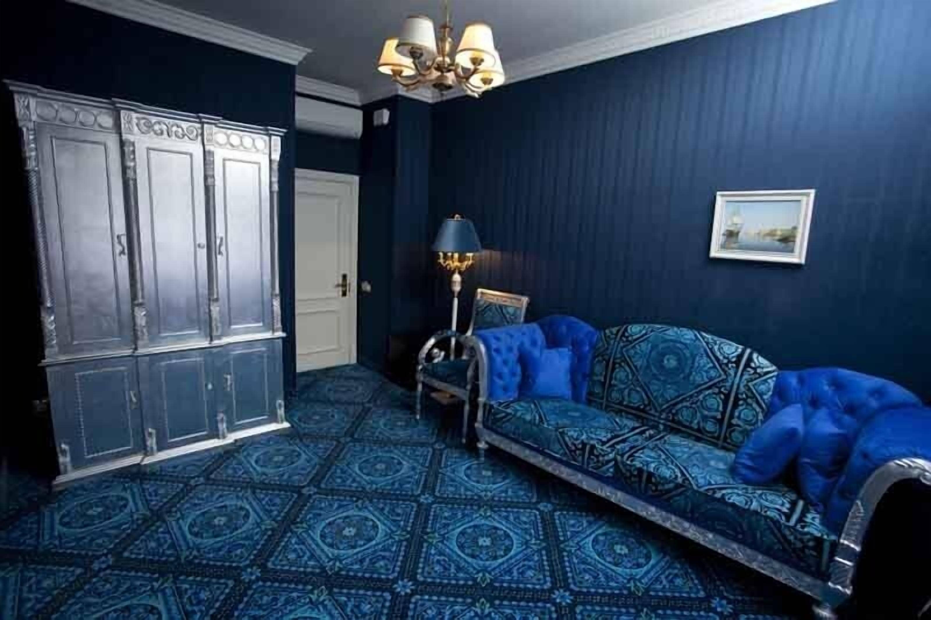 A room with a patterned blue carpet, a blue sofa with a patterned cover, a metal-framed chair, a floor lamp, and a wardrobe with a decorative top.