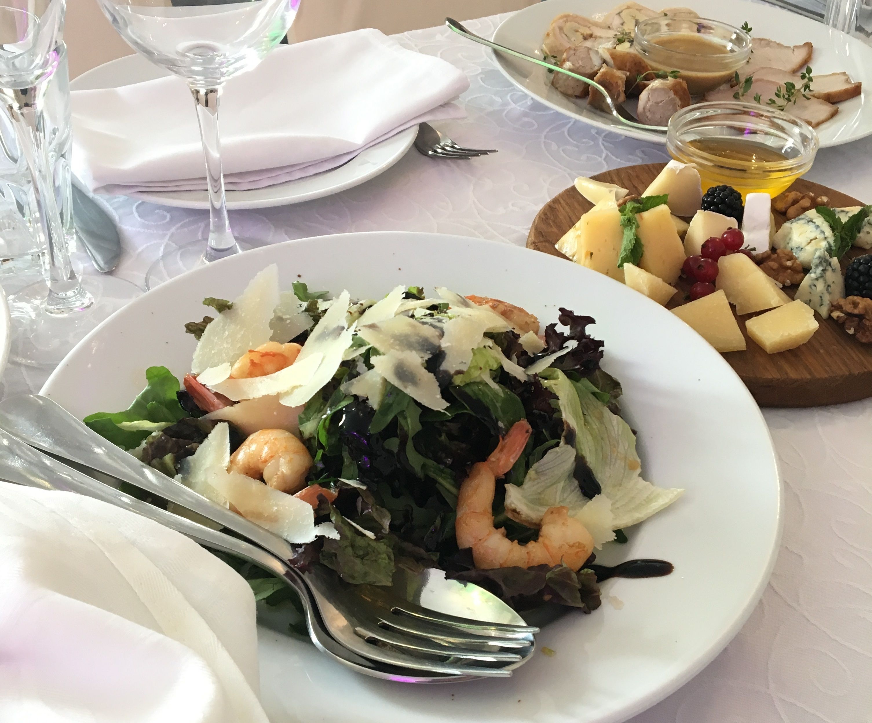 A table set with a plate of shrimp and salad, a wooden board with cheese and nuts, and a glass of wine.