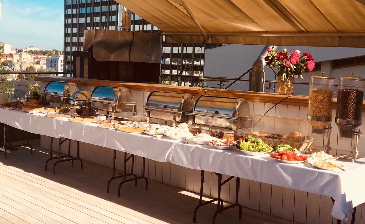 A buffet setup on a rooftop deck with multiple food stations, a large grill, and a flower vase.