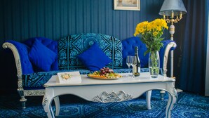 Una habitación con un sofá azul estampado, una mesa blanca con un jarrón de flores amarillas, un plato de fruta y una copa de vino.