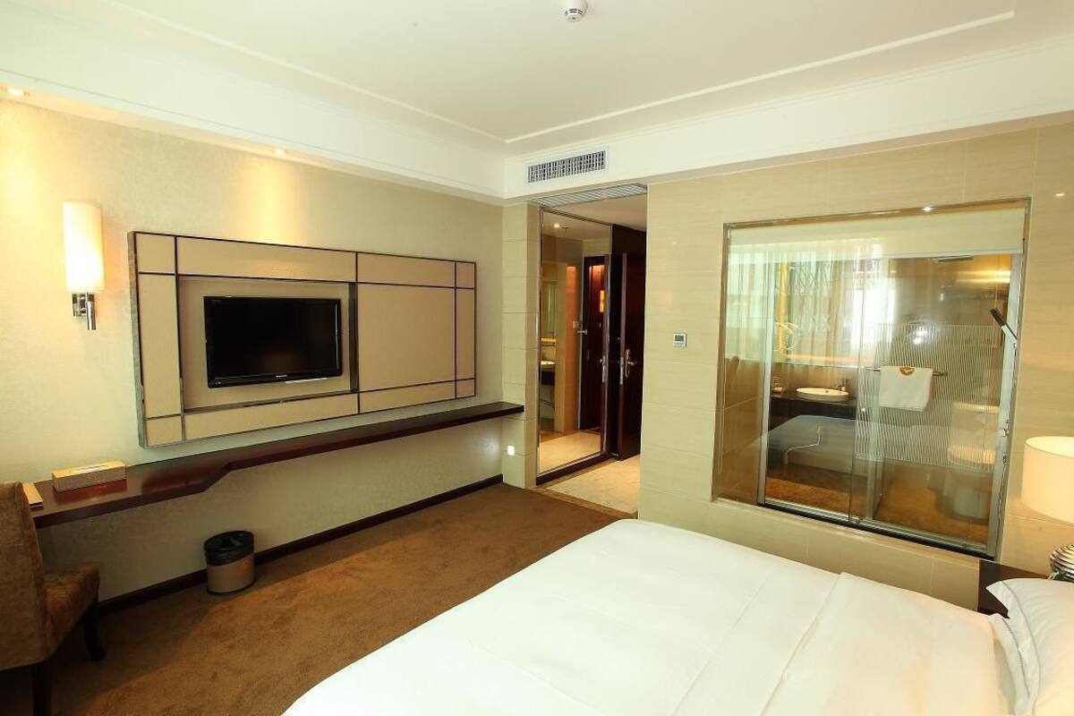 Habitación de hotel con cama, televisor montado en la pared, baño visible a través de un vidrio y un cesto de basura.