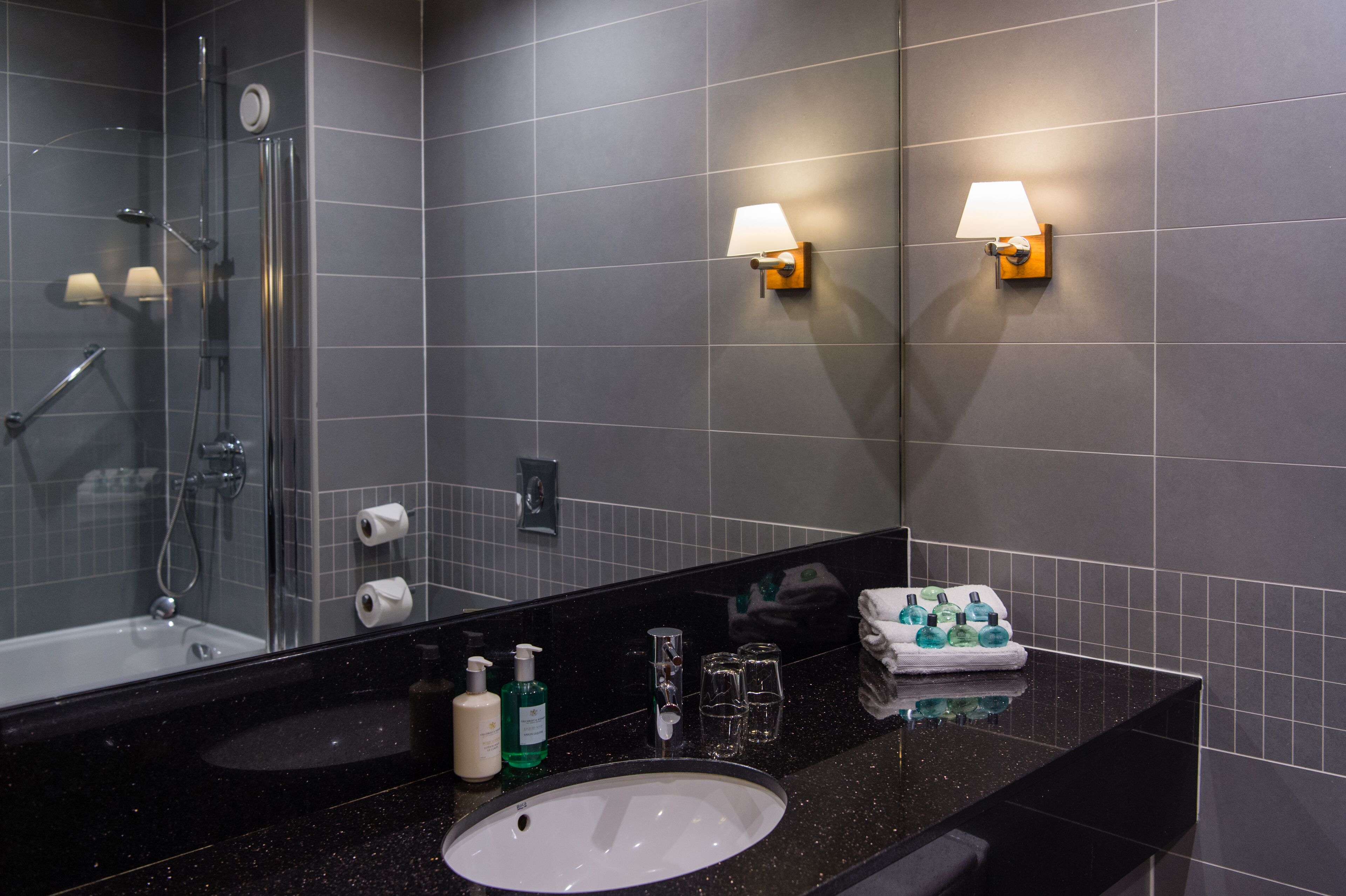 Un baño moderno con encimera negra, lavabo blanco, espejo, ducha y dos luces empotradas.