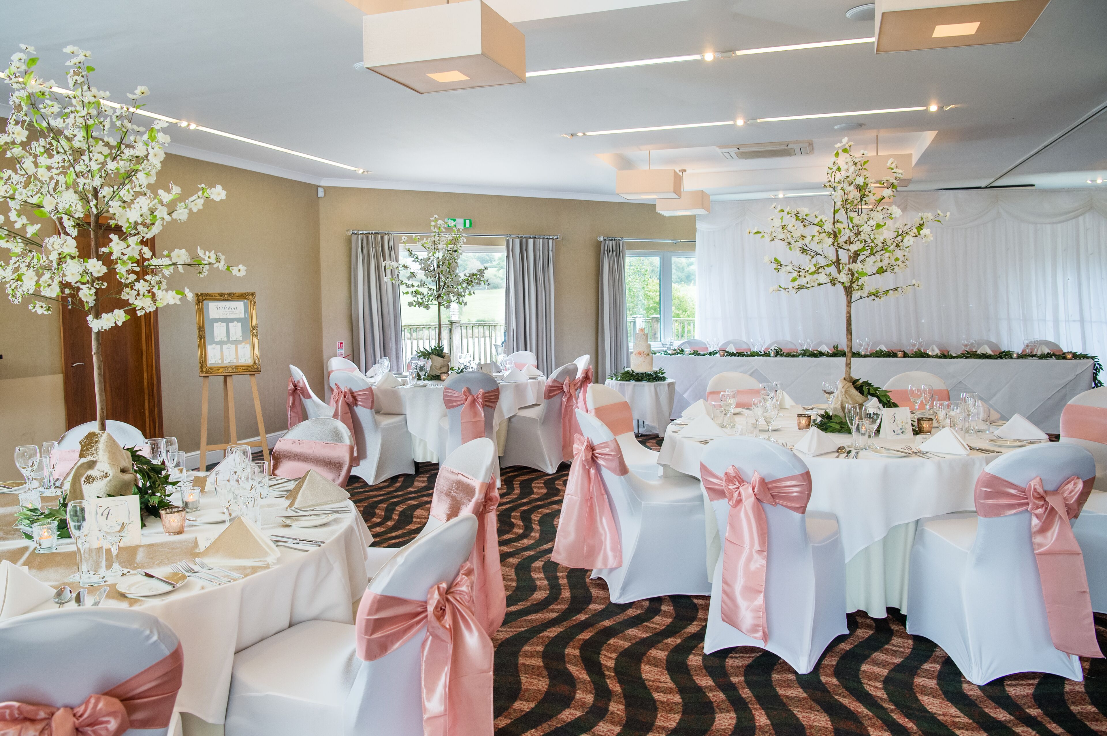 A banquet hall with round tables, white and pink chairs, and floral centerpieces.