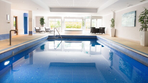 A large indoor swimming pool with clear blue water, surrounded by lounge chairs and a view of greenery outside. - Lakeside Park Hotel & Spa (Ryde)