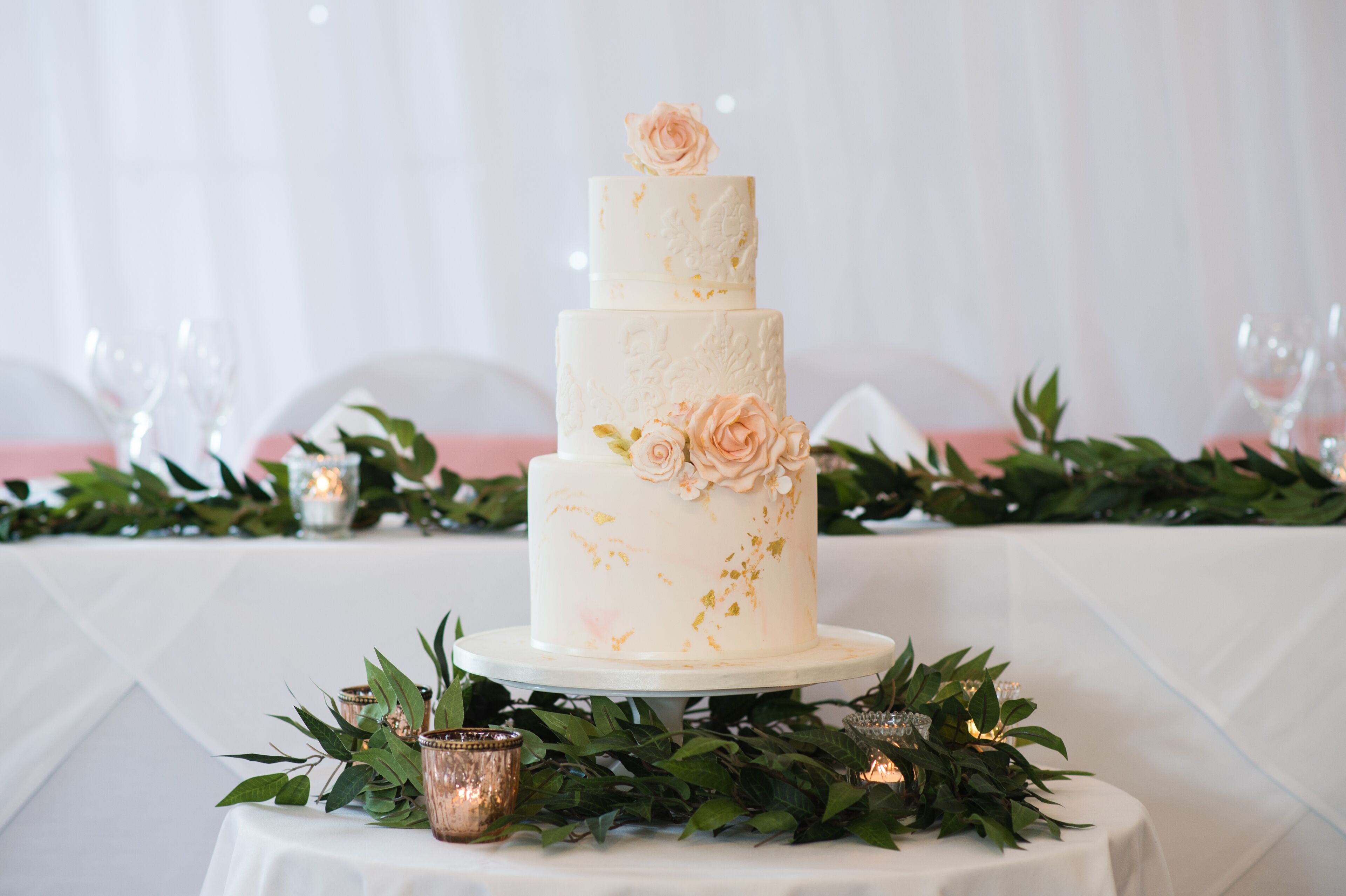 A three-tiered wedding cake with floral decorations on a white tablecloth, surrounded by greenery and candles.