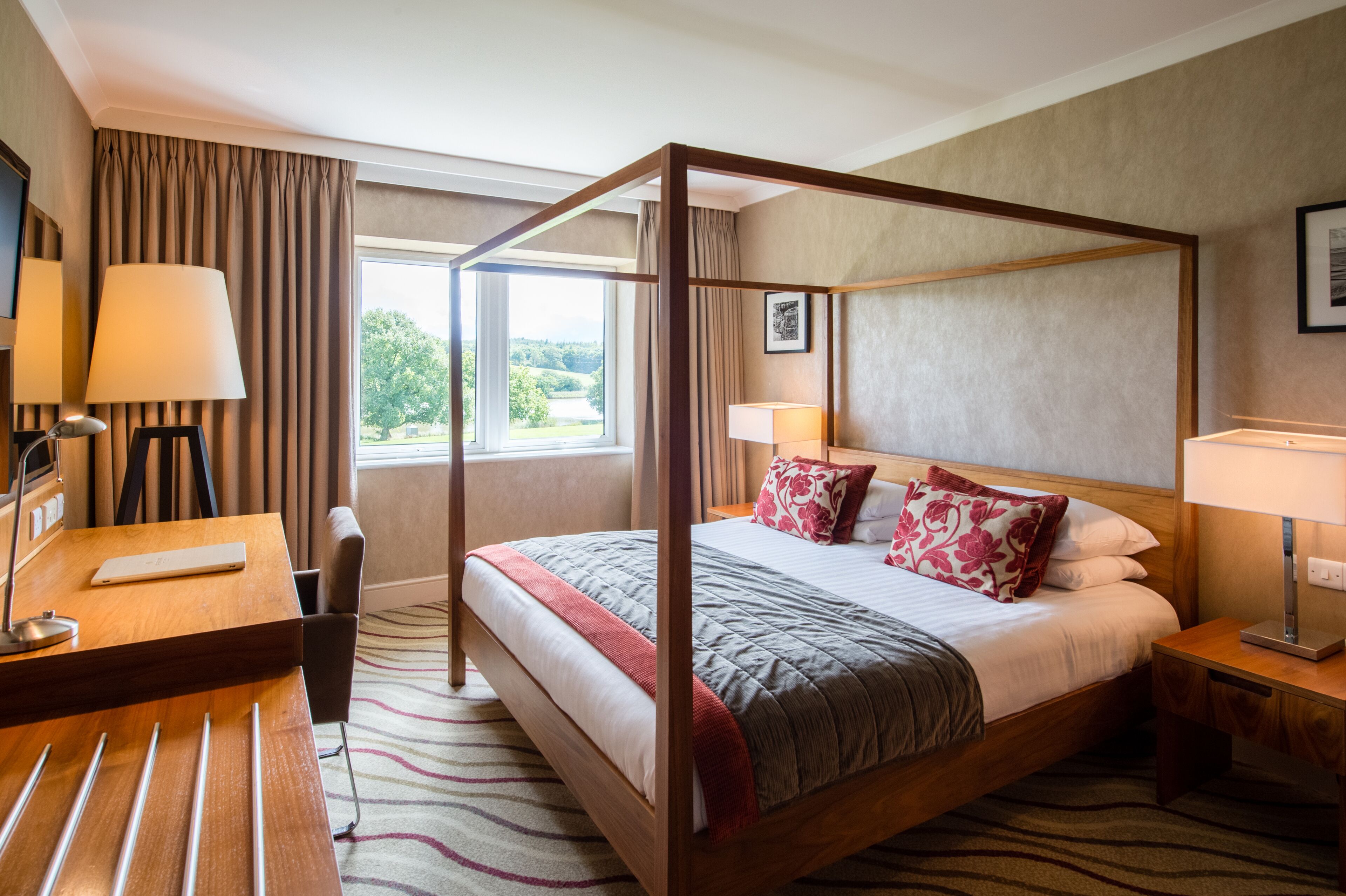 A four-poster bed with a wooden frame, a desk with a lamp, and a view of greenery outside the window.