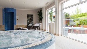 A hot tub with a tiled edge, a seating area with two chairs, and a large window with a view of a pool and greenery. - Lakeside Park Hotel & Spa (Ryde)