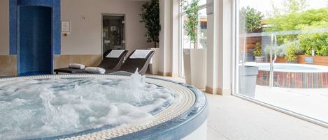 A hot tub with a tiled edge, a seating area with two chairs, and a large window with a view of a pool and greenery.