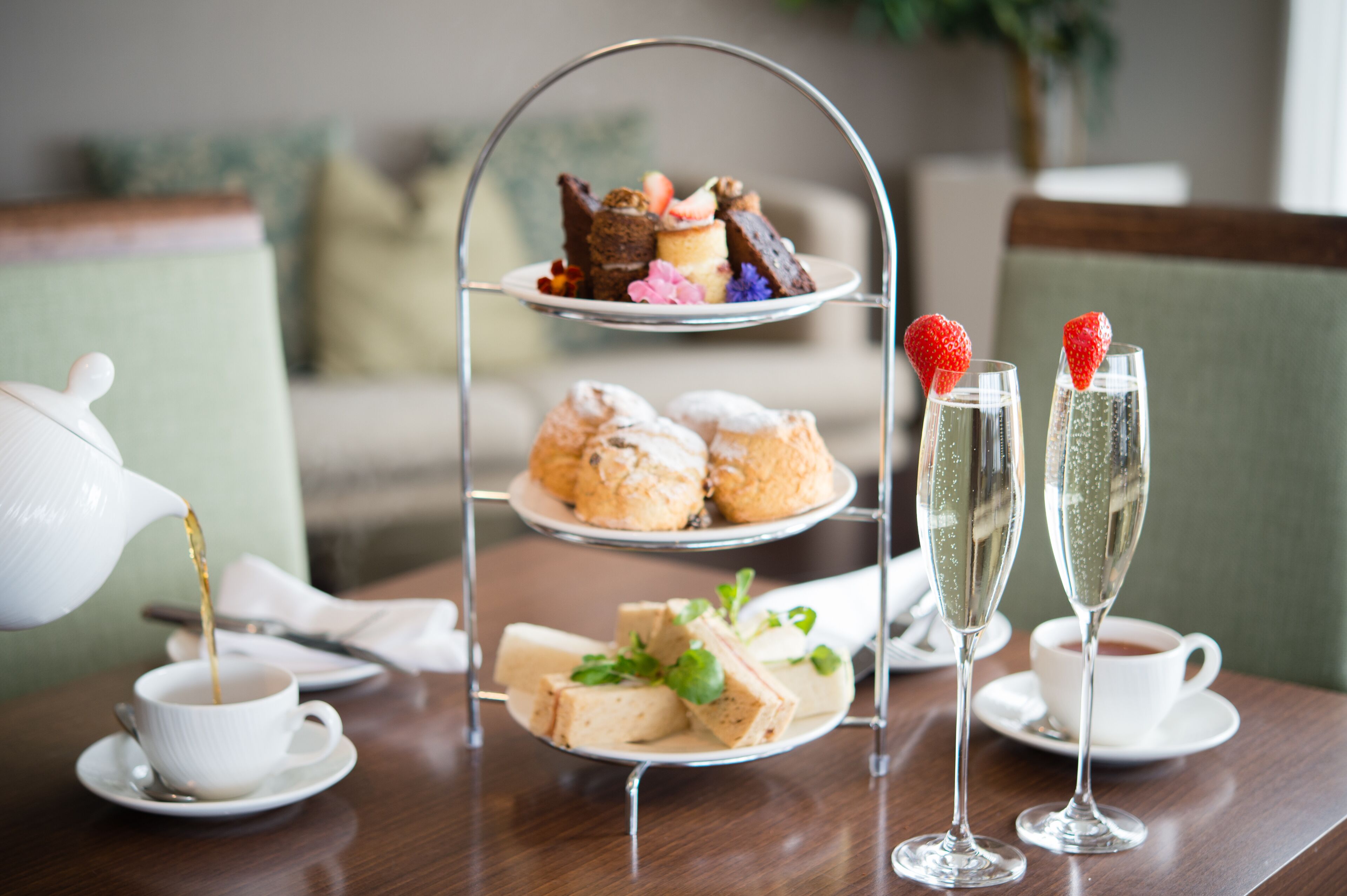 A three-tiered dessert stand with pastries and sandwiches, two champagne flutes with strawberries, a teapot pouring tea, and a cup of tea on a wooden table.