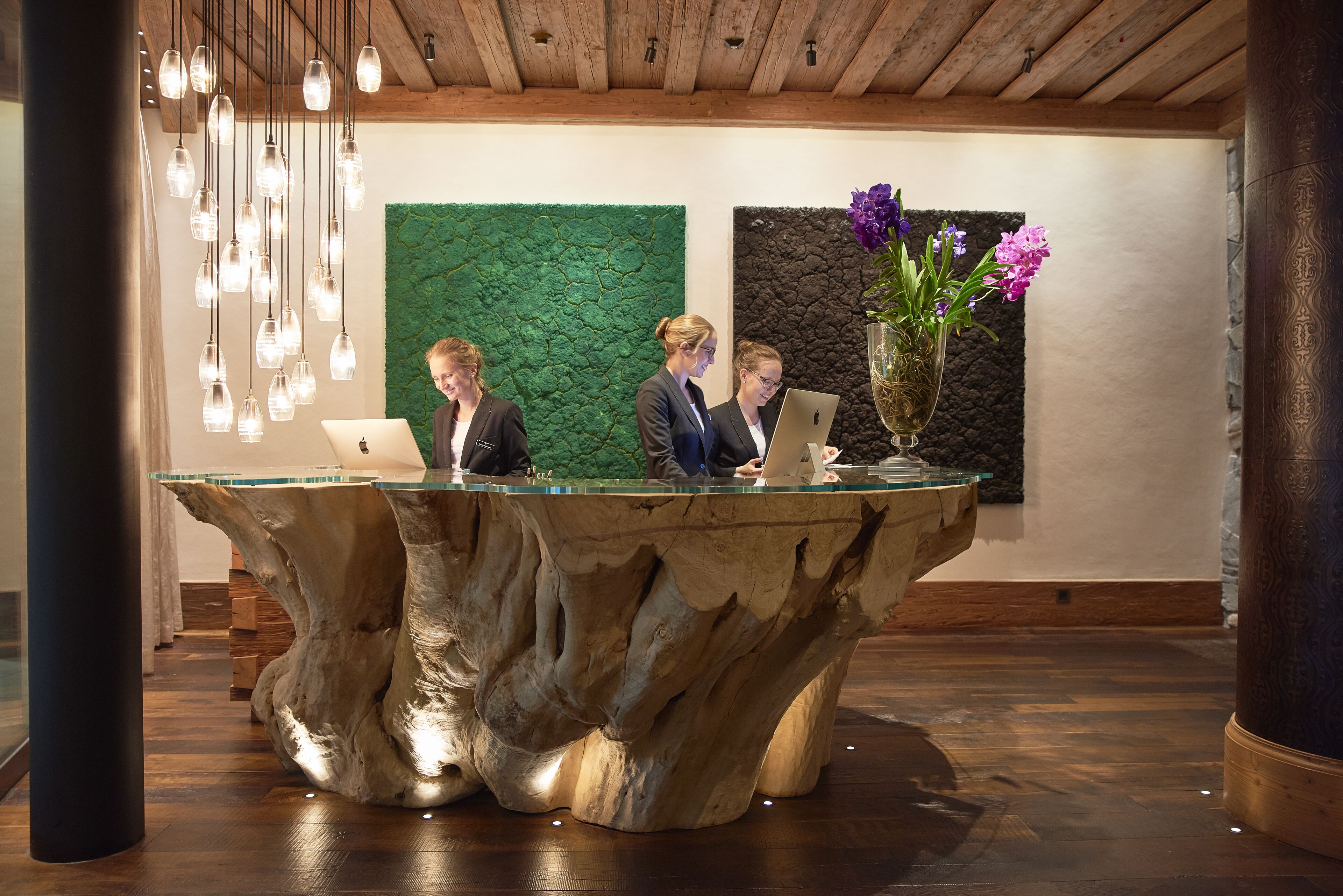 A reception desk with a unique wooden base, a green textured wall, and a vase with purple flowers.
