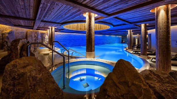 Una piscina interior con techo de madera y paredes de piedra, rodeada de rocas grandes.