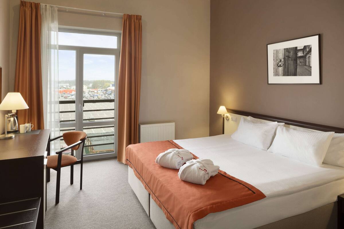 Habitación de hotel con cama, escritorio, silla, ventana con cortinas e imagen enmarcada en la pared.