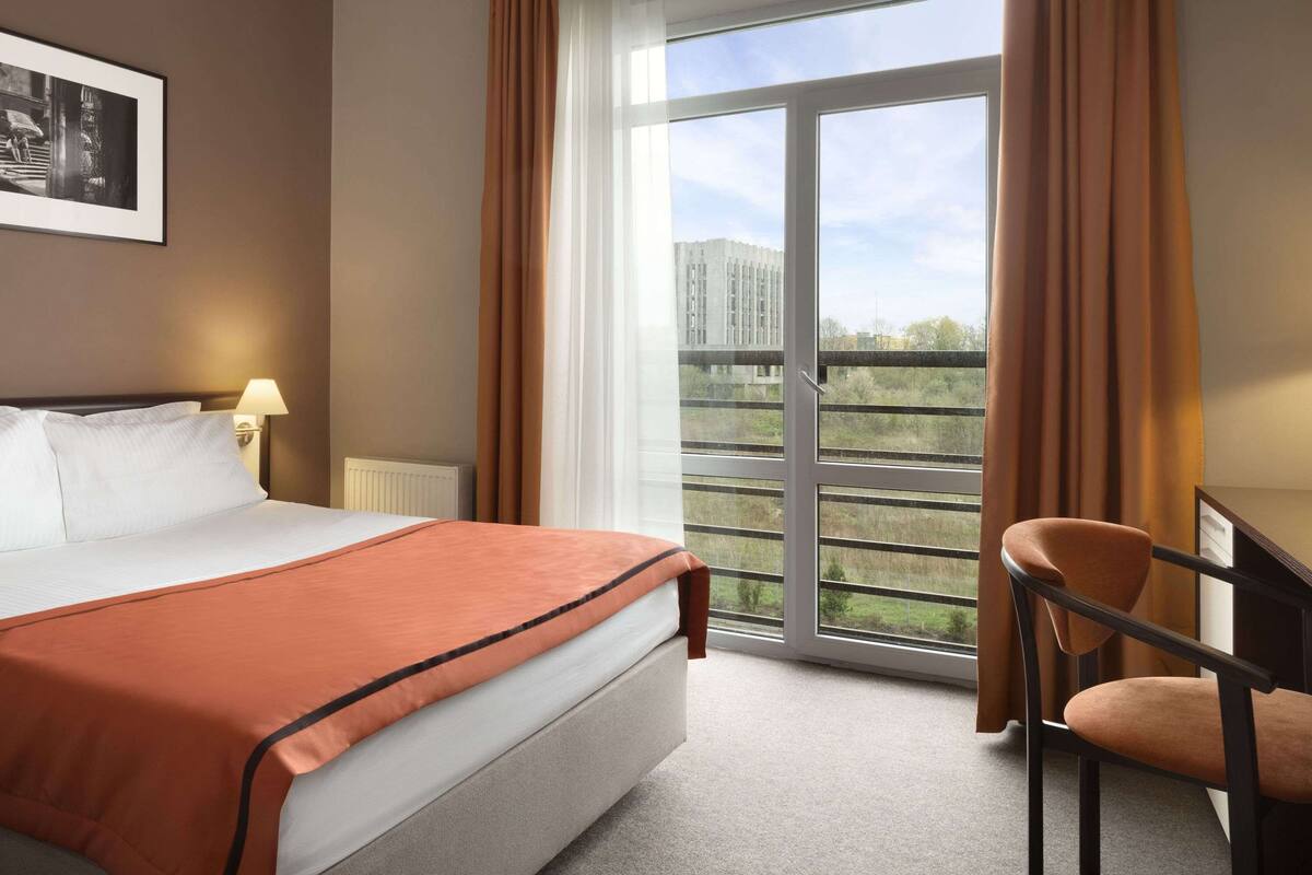 Habitación de hotel con cama, escritorio, silla y un amplio ventanal con vistas a edificios y zonas verdes.