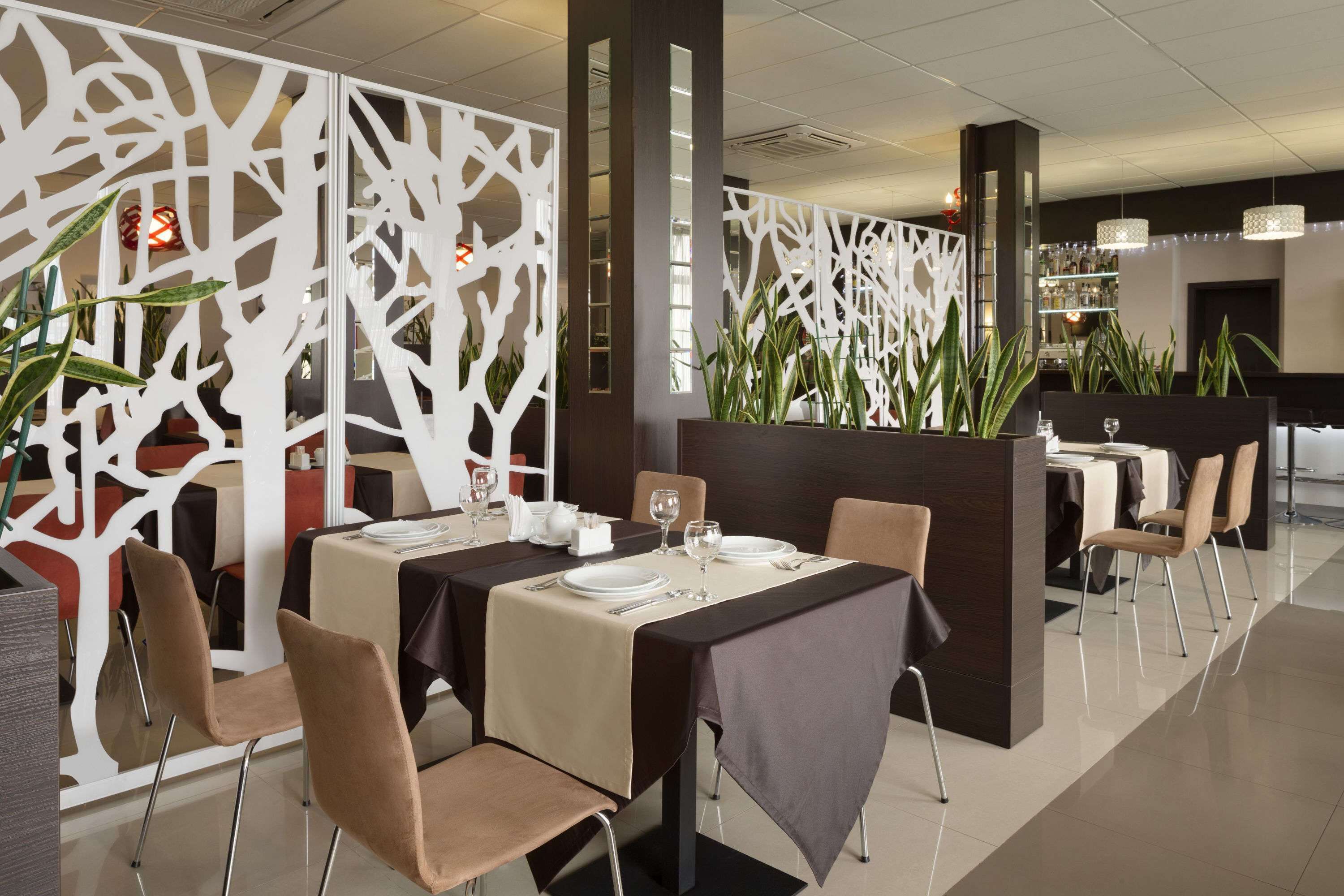 A modern restaurant with a dining area featuring tables set for two, beige and brown chairs, and a decorative white partition with plant arrangements.