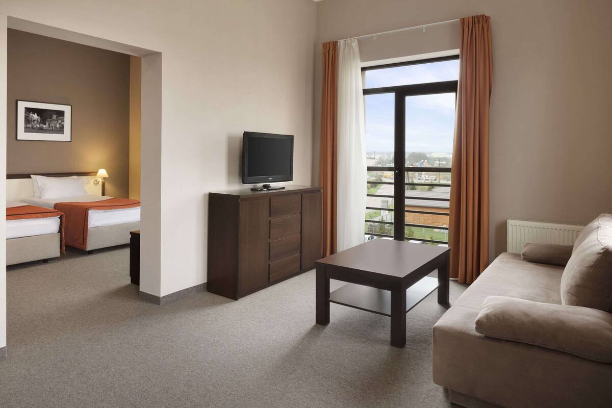 Habitación de hotel con cama, televisor en un mueble, sofá y ventana con cortinas.