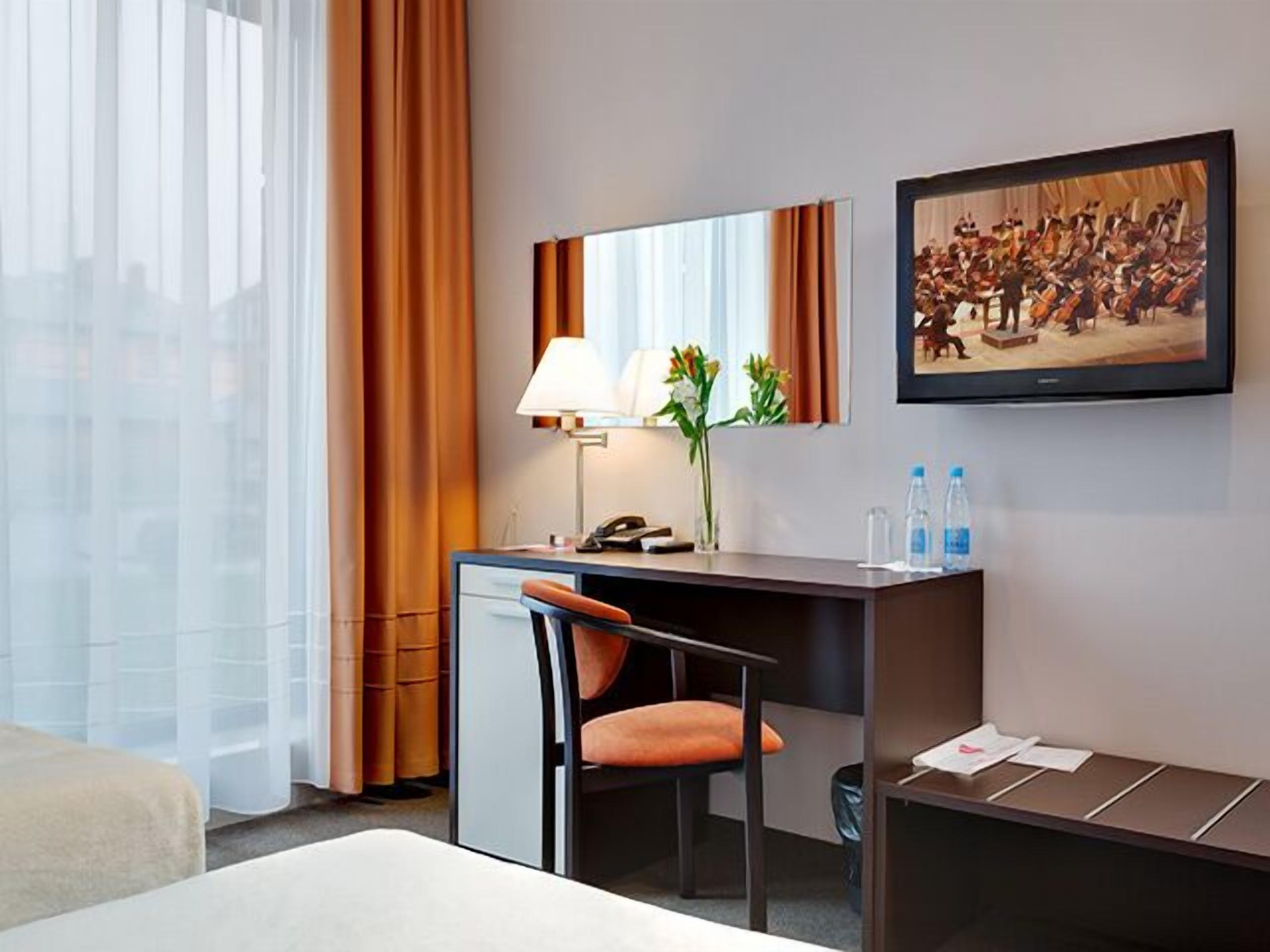 Habitación de hotel con escritorio, silla y un amplio ventanal con cortinas. Hay una lámpara, un florero con flores y un cuadro en la pared.