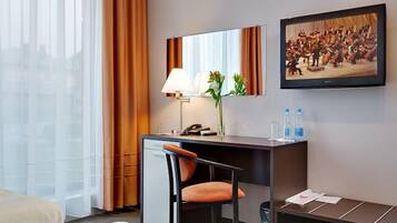 Habitación de hotel con escritorio, silla y un amplio ventanal con cortinas. Hay una lámpara, un florero con flores y un cuadro en la pared.