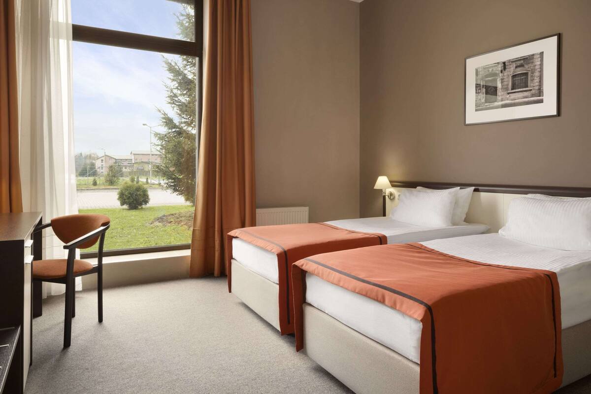 Habitación de hotel con dos camas, un escritorio y una silla. Se ve una ventana con cortinas, una lámpara y un cuadro en la pared.