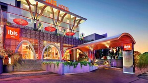 Un hôtel moderne, reconnaissable à son panneau « Ibis Hotels », doté d’une entrée couverte et d’une façade décorative agrémentée d’arcades et de plantes.