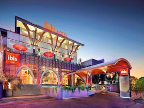 Un hôtel moderne, reconnaissable à son panneau « Ibis Hotels », doté d’une entrée couverte et d’une façade décorative agrémentée d’arcades et de plantes.