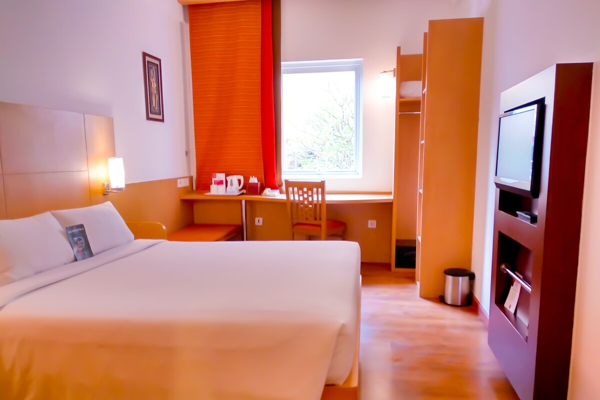 Une chambre d’hôtel comprenant un lit, un bureau avec une chaise, une télévision et une fenêtre avec des stores.