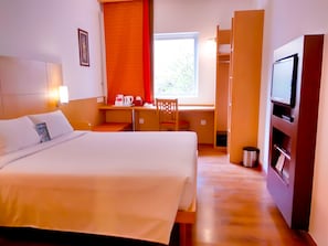 A hotel room with a bed, a desk with a chair, a television, and a window with blinds. - ibis Bali Kuta (Tuban)