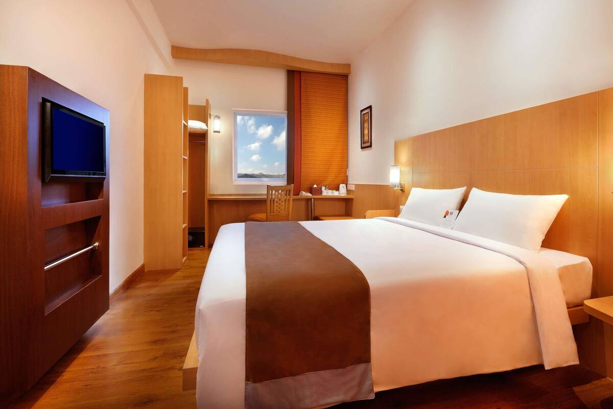 A hotel room with a large bed, a television mounted on a wooden cabinet, a desk with a chair, and a window with a mountain view.