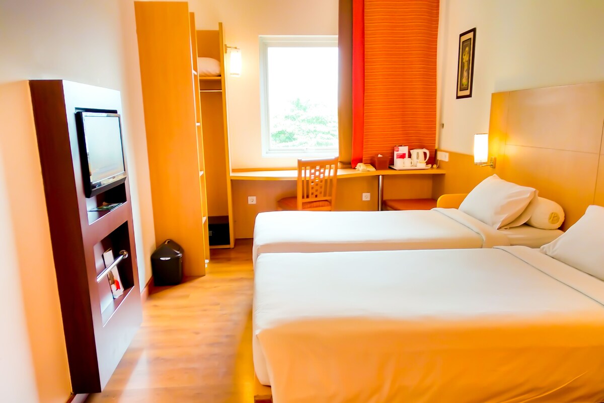 Une chambre d’hôtel avec deux lits, un bureau avec une chaise, une télévision et une fenêtre avec des rideaux.