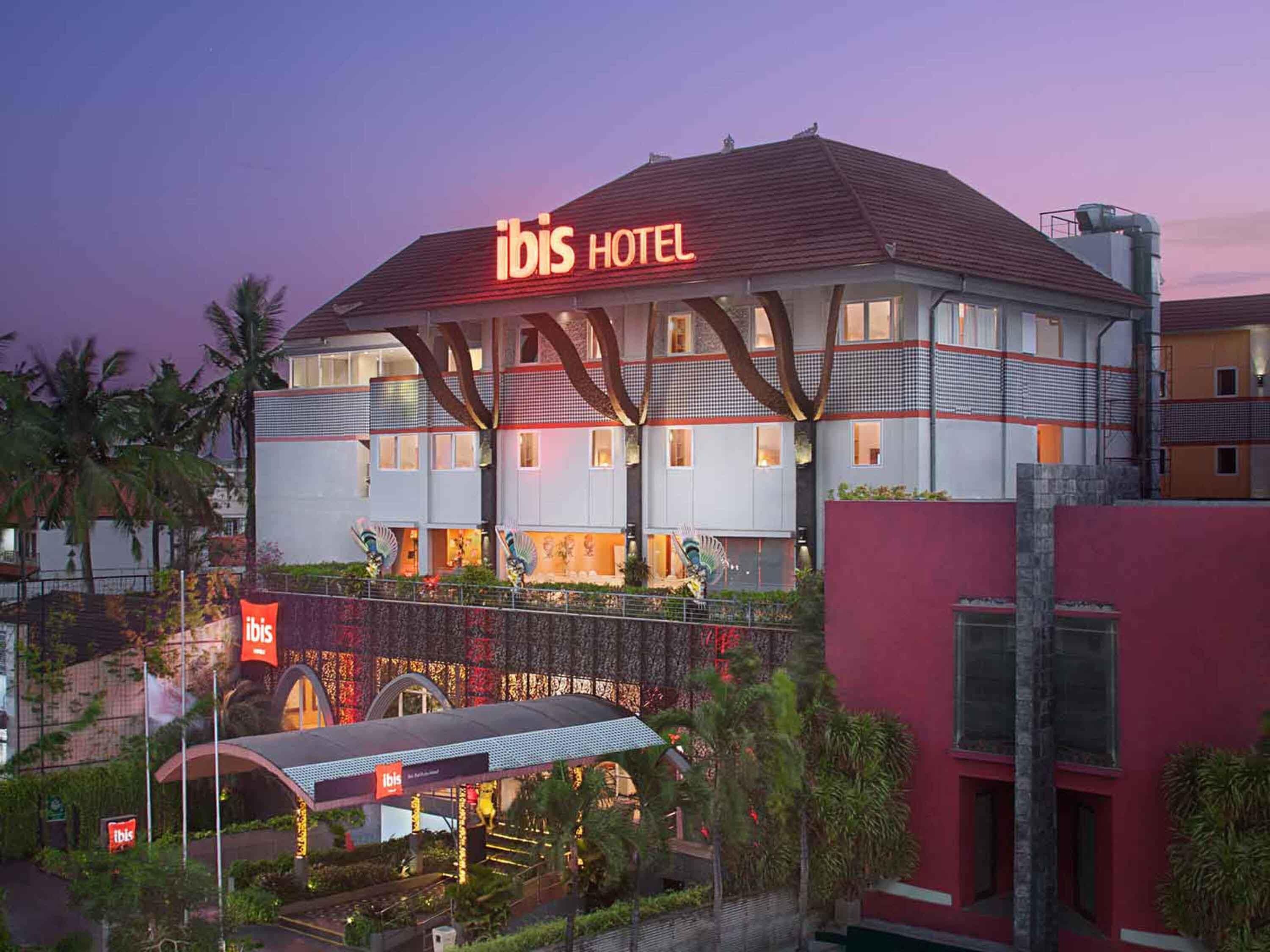 A hotel with a prominent 'ibis HOTEL' sign, a covered entrance, and a landscaped garden area.
