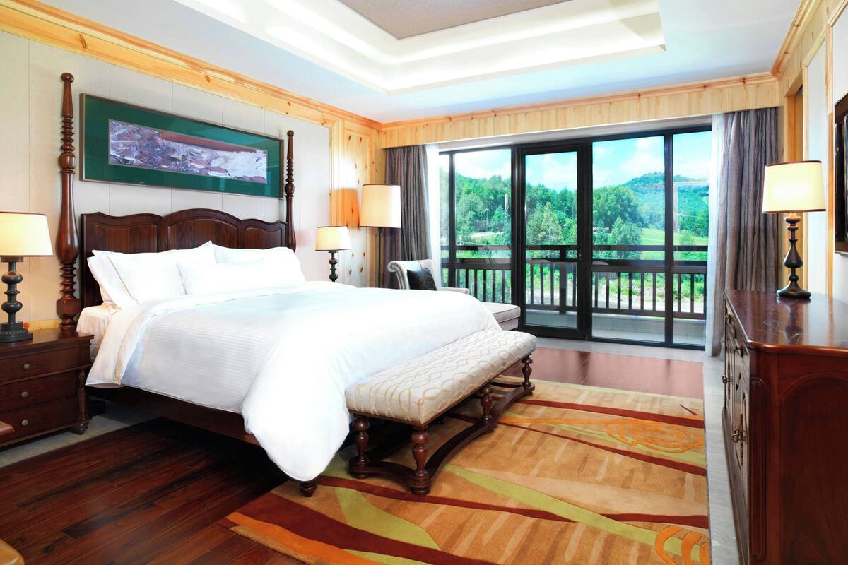 A hotel room with a large bed, a bench, a wooden nightstand, a desk, and a view of a balcony with a scenic landscape.