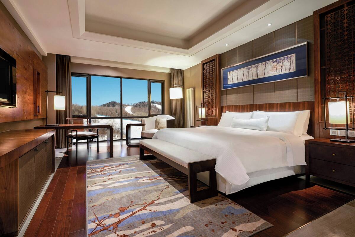 A modern hotel room with a large bed, a desk, and a chair. There is a large window offering a view of the mountains.