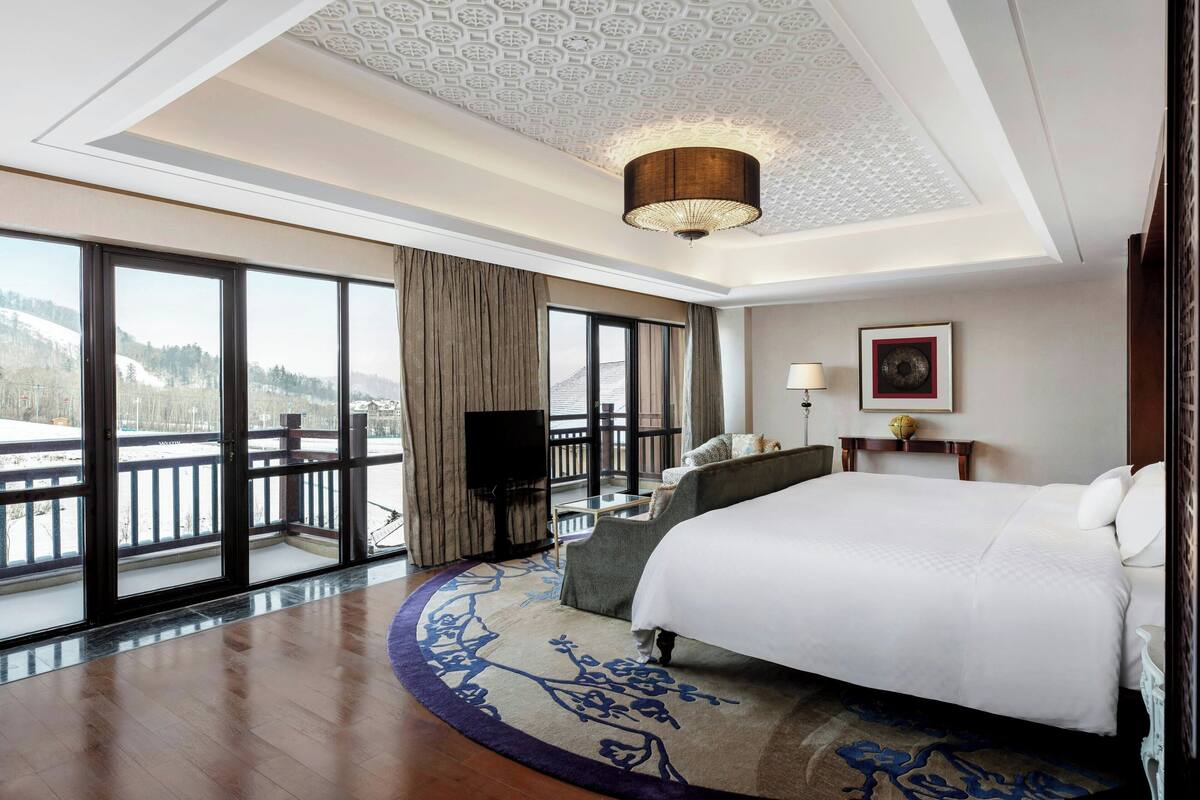 A hotel room with a large bed, a desk, a chair, a TV, and a balcony with a view of the mountains.